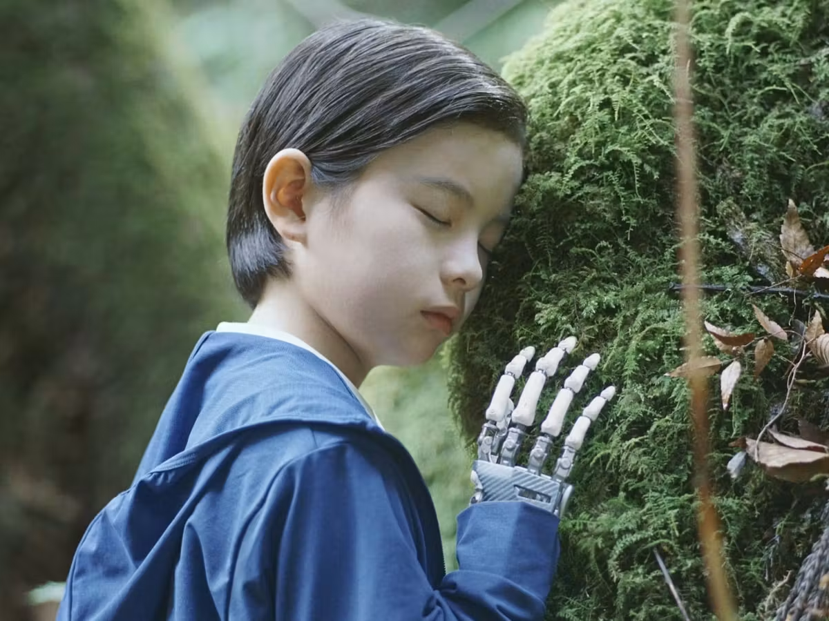 Boy with robot hand leaning meditatively against mossy fern.