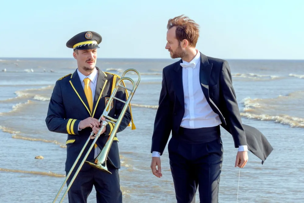 Two men standing on the shore. One in a conductor's tux, the other in a marching band uniform and carrying a trombone.